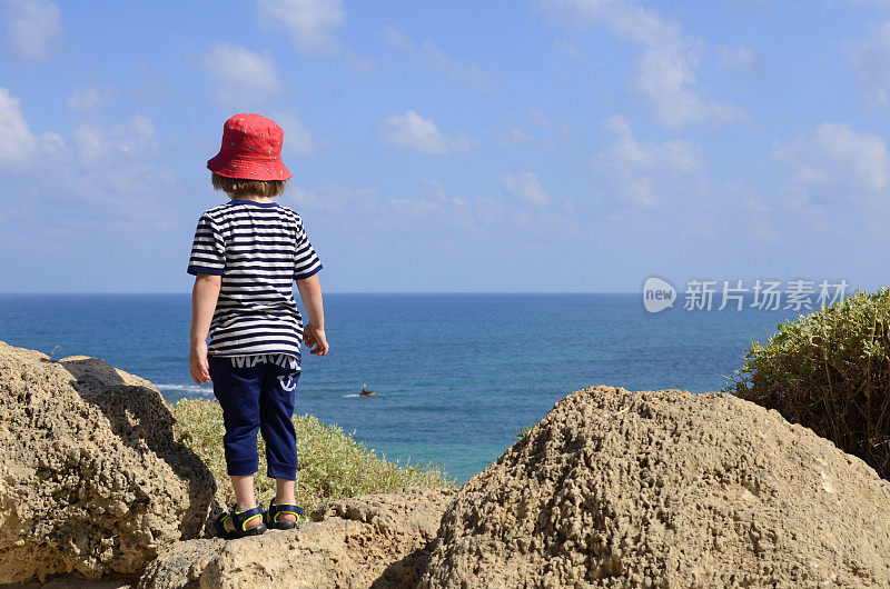 The boy stands with his back and looks at the sea, there is an anchor on the child’s clothes, dreams about a trip, a yacht. Little sailor